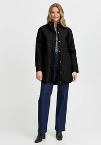 Chaqueta negra acolchada con cuello alto, botones a presión y corte relajado. Combinada con jeans azul oscuro y botas negras hasta el tobillo.
