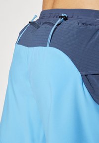 Light blue shorts with a dark blue textured waistband, featuring drawstrings and a pocket detail. Smooth fabric finish, athletic design.