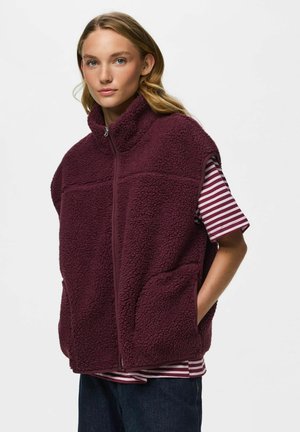 Junge Frau trägt eine weinrote Sherpa-Fleeceweste über einem gestreiften Kurzarmshirt mit den Händen in den Taschen der Weste.