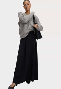 Šedý žebrovaný svetr s dlouhými rukávy, v kombinaci s černou maxi sukní s plisemi a černou kabelkou přes rameno, na neutrálním pozadí.