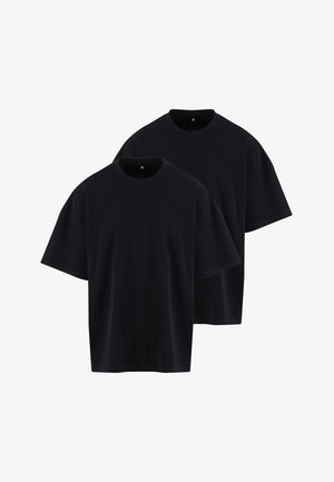 Ung mand iført en løs sort T-shirt og lyse bukser, stående med hænderne i lommerne og kigger let til siden.