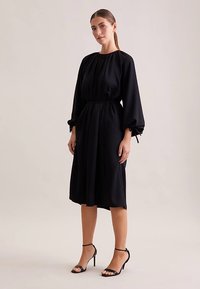 Schwarzes langärmeliges Kleid mit gerafftem Ausschnitt, tailliertem Bund und fließendem Rock; verfügt über weichen Stoff und elastische Puffärmel.
