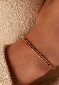 Gouden schakelarmband met in elkaar vergrendelde schakels, met een gladde afwerking en een lichte glans, gedragen op een gestructureerd, beige oppervlak.