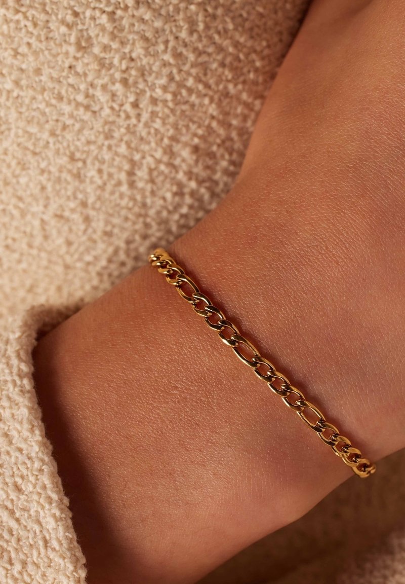 Gouden schakelarmband met in elkaar vergrendelde schakels, met een gladde afwerking en een lichte glans, gedragen op een gestructureerd, beige oppervlak.