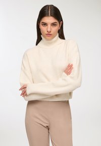 Sweter w kolorze kremowym, teksturowany, z wysokim kołnierzem, szerokimi rękawami i krojem na wysokości bioder, zestawiony z dopasowanymi beżowymi spodniami.