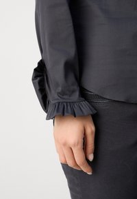 Main reposant sur le côté, portant un haut noir à manches longues avec un poignet à volants et un pantalon noir avec un détail de fermeture éclair visible.