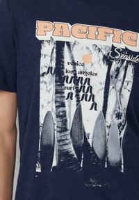 Marineblauwe t-shirt met een grafische print van surfplanken, palmbomen en de tekst "PACIFIC Seaside," met een beige cirkelvormige accent.