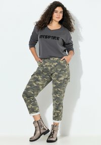 Kobieta z kręconymi włosami, ubrana w ciemnoszary sweatshirt z napisem "INSPIRE", kamuflażowe spodnie cargo i metaliczne buty na sznurówki, stojąca z rękami w kieszeniach.