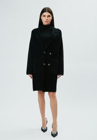 Cappotto nero doppiopetto in tessuto testurizzato, con rever a punta, tasche frontali e bottoni decorativi. Abbinato a scarpe nere con tacco.