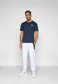 Tengerészkék, rövidujjú póló, mellkasi címkével, fehér sportnadrággal párosítva, oldalsó zsebekkel és texturált felülettel.