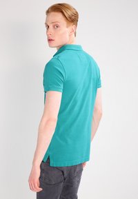 Polo shirt turchese con maniche corte, texture a costine e spacchetti laterali. Presenta un colletto e un design uniforme. Indossato con pantaloni grigi scuri.