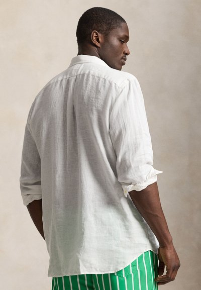 Mężczyzna w białej koszuli z długim rękawem, z podwiniętymi rękawami i zielonych spodniach w białe pionowe paski, stoi tyłem, głowa obrócona na bok.