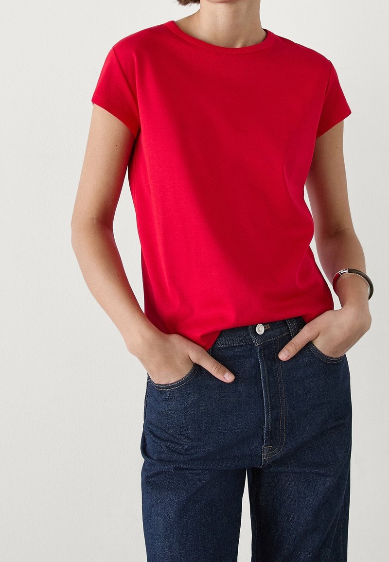 Femme portant un t-shirt rouge uni à manches courtes, rentré dans un jean taille haute bleu foncé, les mains dans les poches, avec un bracelet argenté au poignet droit.