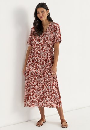 Frau steht in braunem Midikleid mit weißem floralem Paisley-Muster und kurzen Ärmeln, trägt braune Sandalen und goldene Creolen.