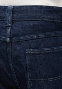 Ζώνη και πίσω τσέπη από σκουρόχρωμο μπλε τζιν με ορατές λευκές ραφές σε λευκό φόντο.