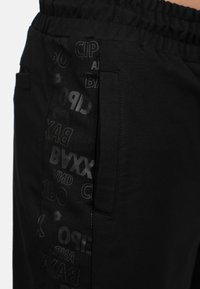 Pantalons de jogging noirs en tissu lisse, avec une taille élastique et un panneau latéral arborant un motif brillant de texte et de logos.