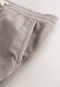 Gros plan d'une poche de pantalon en velours côtelé gris clair avec des rainures verticales visibles et une ceinture élastique sur un fond clair.