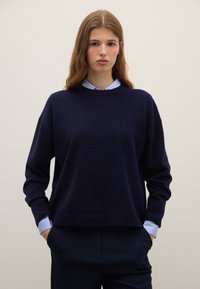 Marineblauer, gestrickter Pullover mit rundem Ausschnitt und überschnittenen Schultern, kombiniert mit einem hellblauen Hemd mit Kragen darunter, mit gerippten Bündchen.