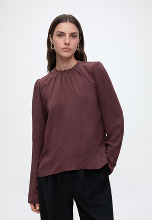 Femme aux cheveux foncés portant des boucles d'oreilles créoles dorées, un haut bordeaux texturé à manches longues avec un col froncé, et un pantalon noir sur fond blanc.