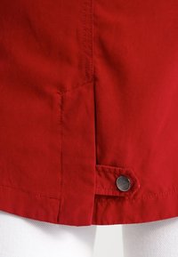 Detalj av röd jacka som visar ett mjukt tyg, en ren fåll och ett metalltryckknappslås nederst till höger. Sömmarna är synliga längs med sömmarna.