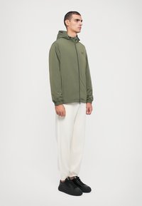 Grønn hettejakke med glidelås foran, elastiske cuffs og en subtil logo, kombinert med hvite bukser og svarte joggesko. Jevn stofftekstur.