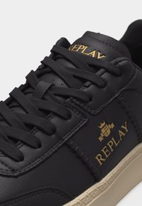 Schwarzer Ledersneaker mit schwarzen Schnürsenkeln, beiger Sohle und goldfarbenem "REPLAY"-Logo auf der Zunge und Seite mit Kronenmotiv.