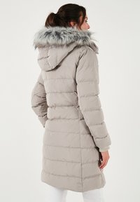Padded dugi kaput u svijetlo sivoj boji s kapuljačom od umjetnog krzna. Ima horizontalno quiltane šavove i bočne džepove. Mekana tekstura i opušteni kroj.