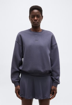 Persoană tânără de culoare, în picioare, purtând un hanorac supradimensionat gri închis și o fustă gri închis asortată, pe un fundal simplu și deschis.