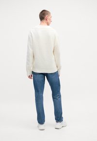 Fet chunky hvit strikkegenser med ribbet hals og nederkant, sammen med blå denimjeans og hvite joggesko. Enkel bakgrunn, person sett bakfra.