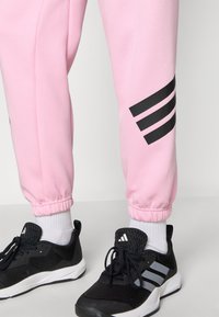 Lyserøde sweatpants med sorte striber, elastiske manchetter og hvide ankelstrømper, parret med sorte sneakers med hvid sål og logo.
