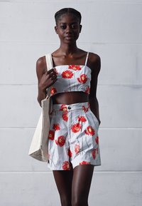 Conjunto de top cortado branco e calções com estampado floral vermelho, apresentando alças finas e bolsos. A modelo segura uma bolsa de mão bege.