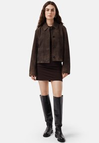 Giacca cropped in suede marrone con colletto, tasche frontali e bottoni neri. Abbinate a un vestito marrone e stivali neri alti e con texture.