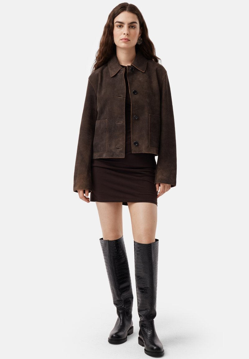 Giacca cropped in suede marrone con colletto, tasche frontali e bottoni neri. Abbinate a un vestito marrone e stivali neri alti e con texture.
