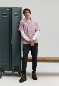 Roze korte mouwen overhemd met knopen over een wit shirt met lange mouwen, gecombineerd met zwarte broek en bruine sneakers, naast een groene kluis.