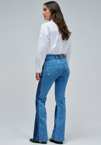 Mujer de pie de perfil vistiendo unos pantalones de mezclilla altos de dos tonos azules, una camiseta blanca de manga larga y zapatos negros de punta, contra un fondo liso.