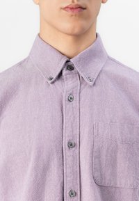 Junger Mann in einem helllila Hemd mit Knopfleiste und Umlegekragen sowie einer Brusttasche, von der Mitte der Brust bis zu den Lippen abgebildet.
