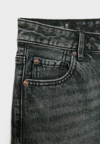 Gros plan sur un jean en denim noir lavé à l'acide montrant la poche avant, la boucle de ceinture et les rivets métalliques avec une étiquette de taille visible à l'intérieur de la ceinture.