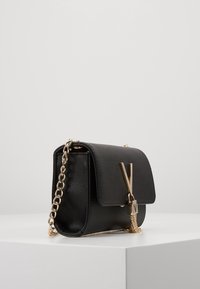 Borsa a tracolla in pelle nera con una catena dorata, dotata di chiusura a patta frontale e di un accento decorativo in metallo a forma di 'V'.