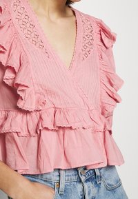 Růžový top s zavinovacím designem, ruffle rukávy a krajkovými detaily, s vertikálním proužkovaným vzorem a vrstveným spodním lemem. Nosený s džíny.