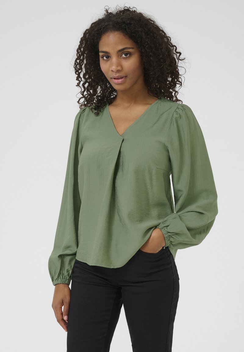 Groene blouse met een V-hals en lange puffmouwen. Gemaakt van gladde stof, met een plooi-detaillering en een elastische manchet. Gecombineerd met een zwarte broek.