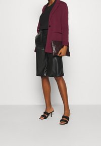 Burgundy kavaj över en svart topp, matchat med svarta läderbyxor. Håller en texturerad mörk handväska. Svarta klacksandaler fulländar outfiten.