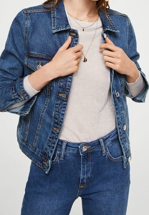 Kvinde iført blå cowboyjakke og jeans over en lysegrå ribbet langærmet skjorte, der holder jakke kraven med begge hænder.
