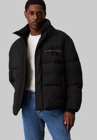 Fekete puffdzseki magas gallérral, elöl cipzárral és hímzett "Calvin Klein Jeans" logóval a mellkason. Tartós anyag, steppelt kialakítással.