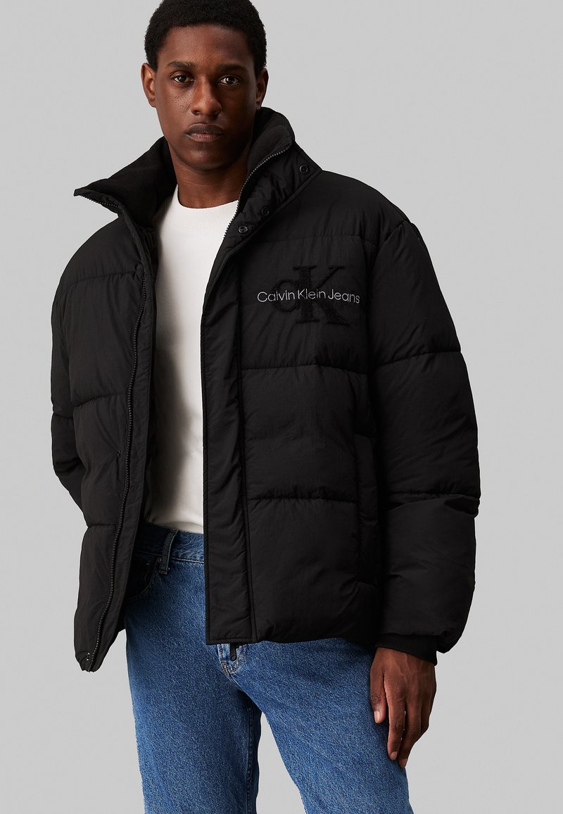 Fekete puffdzseki magas gallérral, elöl cipzárral és hímzett "Calvin Klein Jeans" logóval a mellkason. Tartós anyag, steppelt kialakítással.