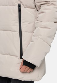 Doudoune beige clair avec un design matelassé, dotée d'une poche à fermeture éclair noire et de poignets noirs côtelés aux manches.