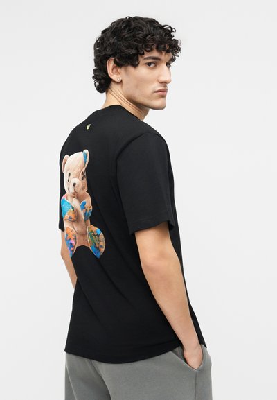 Νέος με σγουρά μαλλιά, φορώντας μαύρο μπλουζάκι με πολύχρωμο σχέδιο αρκουδάκι στην πλάτη, στέκεται με τα χέρια στις τσέπες.