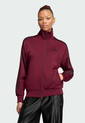 Junge Person trägt eine dunkelrote Adidas Trainingsjacke mit schwarzen Streifen und schwarze Trainingshose, steht mit einer Hand in der Tasche.