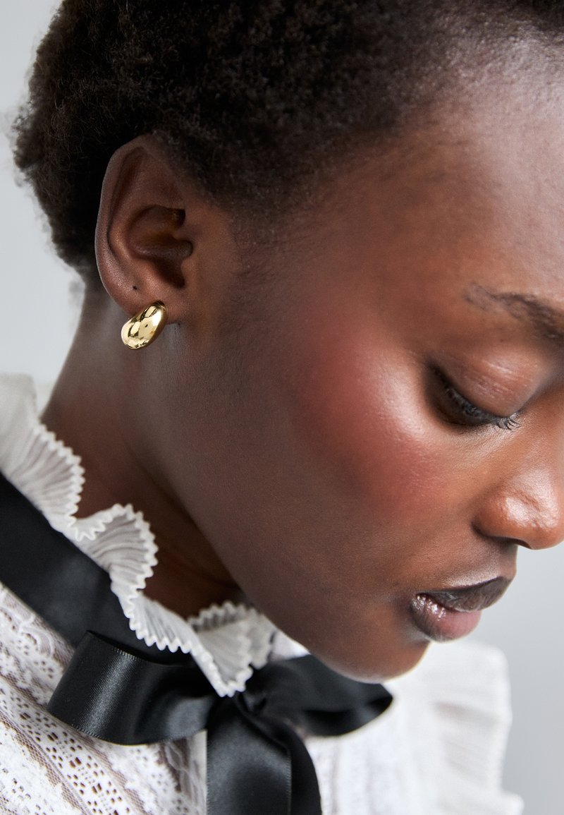 Gul stud ørering i en rund form, glat tekstur, båret af en model med en struktureret frisure. Detaljeret hvid bluse med rynker og sort bånd.