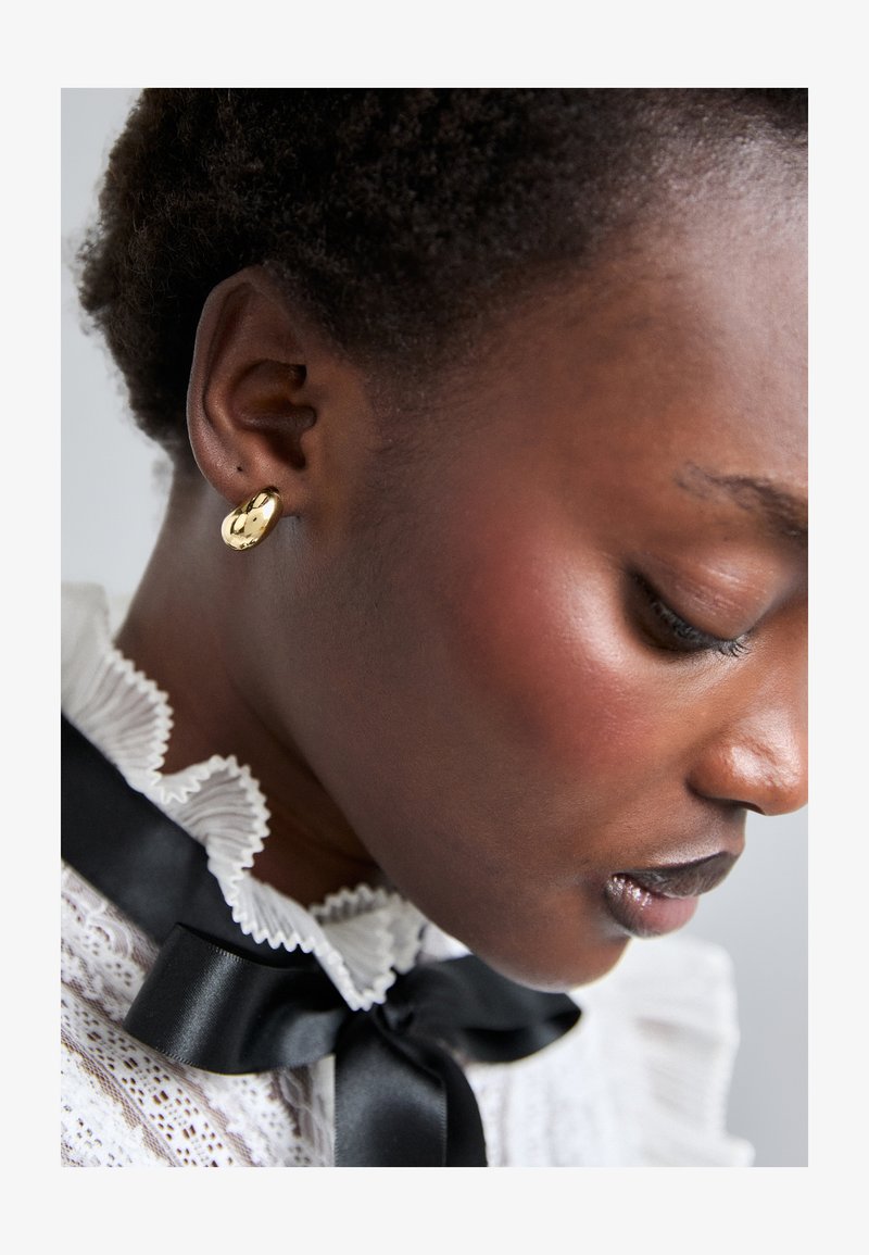 Gul stud ørering i en rund form, glat tekstur, båret af en model med en struktureret frisure. Detaljeret hvid bluse med rynker og sort bånd.