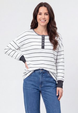 Femme souriante aux longs cheveux bruns portant une chemise blanche à manches longues avec des rayures noires et un jean bleu, debout avec une main sur la hanche.